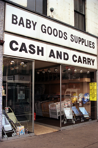 A terraced shopfront with full-length glass windows and a recessed entrance in the middle. Cots and pushchairs are displayed in the windows, and a sign above reads “Baby Goods Supplies / Cash And Carry”.