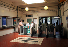 The interior of a small station ticket hall, with a British Rail symbol mosaic on the tiled floor. Three people are standing together at the left of the photo, on different sides of the ticket gates. Globe lights hang from the ceiling. The ticket window in the wall to the left of the ticket gates has the shutter pulled down. Daylight is visible through windows beyond the ticket gates.