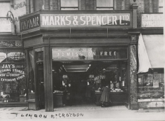 A black-and-white photo showing a small shopfront with signs above it reading “Bazaar”, “Marks & Spencer Ltd”, and “Admission Free”. A few shoppers and shop assistants are looking out of the open shopfront towards the camera. The shopfront protrudes slightly from the shop to its left, giving a diagonal aspect to the side of the shop. Writing is visible on the window of the shop to the left: “Jay's Furnishing Stores Ltd. Cash or credit. Extensive stock.”