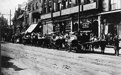 A black-and-white photo of a wide terraced shopfront with several horse-drawn vehicles along the roadside in front.