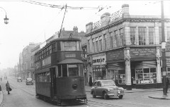 A black-and-white photo showing an old-fashioned overhead-powered tram in the middle of a street.  A corner building on the right has frontages for John Hood Ltd dyers and cleaners, and the Ministry of Labour & National Service Combined Recruiting Centre and Medical Boards.