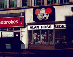 Two neighbouring shopfronts viewed in late-afternoon sunlight with heavy shadows. The left one has a frontage sign (partially cut off) reading “Ladbrokes” and the right one has a sign reading “Alan Ross Sport[s?]”. Another sign above the Alan Ross one shows a drawing of five balls — basketball (or possibly old-style football), tennis, cricket, golf, and football — arranged like the Olympic rings, with three above and two below. This shopfront has shoes and clothes visible in the window, covered by a protective metal mesh.