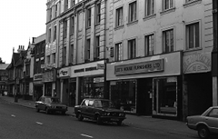 A black-and-white photo of an oblique view onto a row of shops of varying architectural styles. The bulk of the picture is taken up by a tall building in the foreground. The closest shop has a sign reading “Lee’s House Furnishers Ltd”. An opening next to this leads into an alley with a bas-relief sign above it reading “REMOVALS” on a curved baseline over a simple abstract design.