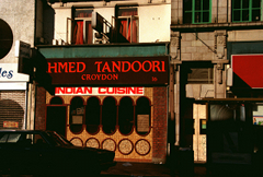 A small restaurant frontage in the middle of a terrace. Much of the image is in shadow. A sign above the restaurant reads “Ahmed Tandoori / Croydon” in orange letters on a brown background, and below it a banner reads “Indian Cuisine” in the same orange on a white background. The front of the restaurant is tiled with small light brown tiles, and a window is set into it formed of five tall narrow ovals attached at their long sides. Five circles along the base of the wall line up with the curves of the ovals.