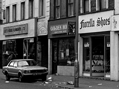 A black-and-white photo of three terraced shopfronts. A car is parked outside one of the shops, and the pavement is badly littered.