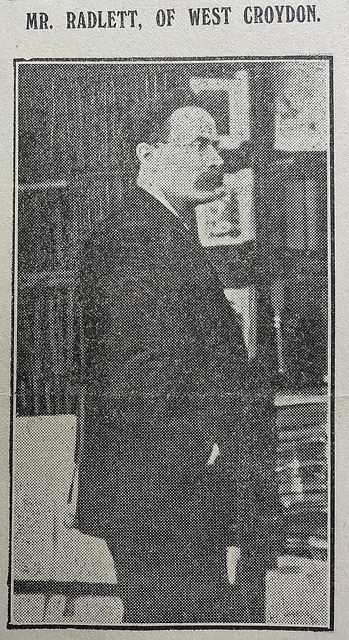 A black-and-white photo of a white man with a moustache, dark hair, and a receding hairline.  He is wearing a dark coat and standing side-on to the camera.  Shelves of books can be seen in the background.