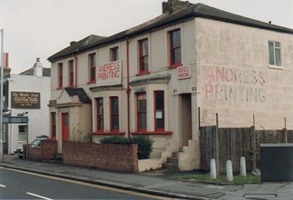 A two-storey building with three pairs of windows along the upper floor. The outside has been painted white, but there are clear signs of weathering. The windowframes and doors are painted red. Two signs on the frontage read “Andress Printing”. A painted sign on the side of the building shows the same text, but has worn away with time. The side of the Wandle Arms pub is visible to the left.