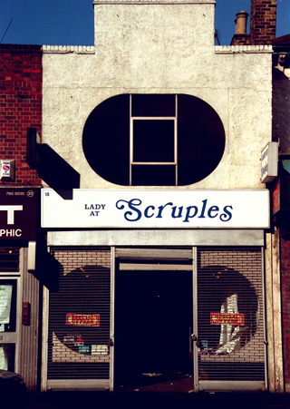 A narrow terraced two-storey shopfront. A striking oval window is set into the first floor, and the roof profile features a large sticking-up rectangular detail. The shopfront on the ground floor has an open door-shaped space in the middle, with grid-style shutters drawn down on either side. A sign above reads “Lady at Scruples”, with the “Lady at” part in smaller text than the “Scruples”.