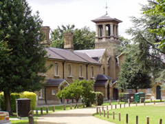 A light-coloured brick building with gabled porches and a belltower with a weathercock on top. Trees of various types grow around, and a wide footpath curves from the viewer towards the building, with grassy stretches on either side.  A number of wheeliebins are somewhat incongruously visible.