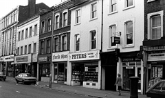 A black-and-white photo of a terrace of shops. In the middle is one with a sign reading “Peters Master Bakers”.