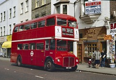 A red double decker bus alongside a three-storey terrace with shops and restaurants on the ground floor.  To the side of the bus is a brick-fronted restaurant with multi-paned windows and a sign reading “Pino’s Place”.