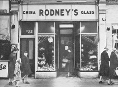 A black-and-white photo tightly focused on a small shopfront with a central recessed doorway and windows on either side.  Several people are walking past and one is standing in the doorway.  A sign above reads “Rodney’s” in the centre with “China” and “Glass” to either side.