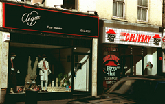 Two terraced shopfronts next to each other. The left-hand one has a sign above reading “Clique” and in smaller text “Pour Homme”, in silver writing on a black background with a thin red border.  In the shop window are three mannequins.  The right-hand one has a sign above with two Pizza Hut logos at each side and the word “Delivery” in the middle.