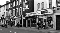 A black-and-white photo of a terraced street, with shopfronts all along the ground level including Quality Food Centre, Fingertips, Croydon Fruiterers, and N B Patel & Sons newsagent.  A few people are walking down the street and standing in front of the newsagent.  A large cigarette advert is visible on a side wall.