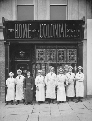 A black-and-white photo of a small shop with 'The Home & Colonial Stores' in metallic letters on a black frontage.  Eight men are standing outside; seven of them in white overalls and one in dark overalls.