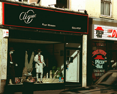 A terraced shopfront with a sign above reading “Clique” and in smaller text “Pour Homme”, in silver writing on a black background with a thin red border.  In the shop window are three mannequins.  The left-hand one is standing up with a dark suit and hat on.  The middle one is upside down and wearing a light greyish suit.  The right-hand one is wearing a light-coloured skirt suit and a hat similar to the one on the first mannequin.  To the right of this shopfront, part of a Pizza Hut shopfront is visible.