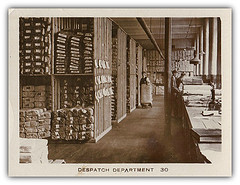A landscape-oriented card showing a sepia photo of a wide corridor in a warehouse.  To the left are shelves stacked with wrapped bundles, twice the height of the person standing next to them.  To the right are two more people working with folded fabric on a wide long workbench.  Below the photo is printed “DESPATCH DEPARTMENT 30”.