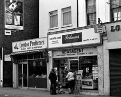 A black-and-white photo showing two small shopfronts: Croydon Fruiterers (“Quality Fruits & Vegetables”) on the left and N B Patel & Sons tobacconist, confectioner, and newsagent on the right. The frontage of Patels bears the words “Rothmans King Size” in large letters.  The wall to the left of Croydon Fruiterers has a large advert for Embassy Number 1 cigarettes.