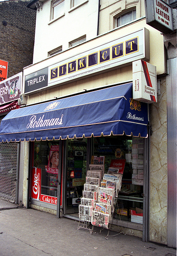 A small terraced shopfront with a rack of newspapers on the pavement just outside. Decals on the windows advertise Coke, a blue canopy advertises Rothmans cigarettes, a projecting sign advertises Embassy Number 1 cigarettes, and a sign above the frontage advertises Silk Cut cigarettes.