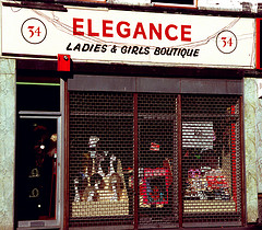 A terraced shopfront with a mesh-style shutter down over the front.  The sign above the frontage reads “Elegance Ladies & Girls Boutique”.  Through the mesh, some mannequin heads with wigs on are visible.