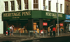 A corner view of a double shopfront with a sign above the frontage reading “HERITAGE PINE” in gold letters on a dark green background.  Pine furniture is visible in the windows, as are signs reading “SALE” in white letters on a red background.  A few people are walking past the shop along the pavement.  The corner of the pavement is separated from the road by metal railings, on which is a sign reading “Oakfield Road”.