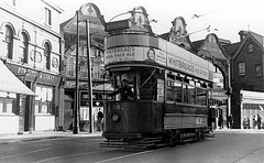 An old-fashioned tram running in front of a parade of shops.  On the left is a frontage reading “Bowditch & Grant”, and to the right of this is another reading “The Creamery” with “Luncheons / Teas” below.  The tram itself carries advertising for Whitbread’s ale, stout, and IPA.