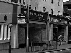 A black-and-white photo of a terrace of shops.  The one in the middle has a sign reading “MA[something]BRO FASHIONS” and whitewashed windows.  A rather fashionable-looking person is walking past the next-door shop (House of Ceramics).