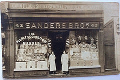 A black and white photo of a terraced shopfront with a fully glazed frontage and two people in overalls standing on either side of the recessed central doorway.  Signs above the shopfront read: “Corn Flour Seed & General Produce Merchant / 43 Sanders Bros 43”.  Writing on the left-hand window reads: “The peoples corn & flour market”.