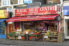 A terraced shopfront with a display of colourful vegetables at the front.  Signs above read “Wholesale & Retail Meat & Poultry” and “Halal Meat Centre / Fresh Beef * Lamb * Poultry * Fish”.