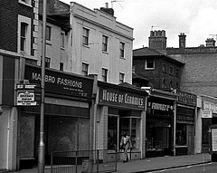 A black-and-white photo showing four shopfronts: Ma[...]bro Fashions, House of Ceramics, Fininley Ltd, and Maloneys.  House of Ceramics has an unusual shopfront with patterned tiles surrounding the windows.  A pedestrian is walking past the front of this shop.