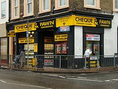 A corner shopfront property with signs for “Albemarle & Bond / Cheque & Pawn / Cheques Cashed / Pawnbroker”.  Railings separate the pavement from the street, and a sign on the side of these reads “Oakfield Road”.