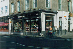 A corner property viewed from the opposite corner of the crossroads.  The frontage on the ground floor is painted black and has a sign reading “Bowman” along with street numbers 60 and 62.  The windows are filled with various items.  Three people are walking past the shop.  Metal fencing runs along the road side of the pavement, and Belisha beacons stand on both sides of the shop.