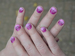 A light-skinned person’s hands held crossed one over the other.  The fingernails are short and have been painted with flame-like designs in pink and magenta.