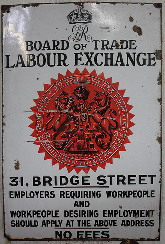 A rectangular metal sign, portrait orientation, painted white with a large circular red “seal” in the middle and a stylised crown with the monogram “GVR” [George V Rex] at the top.  Words in black letters read: “Board of Trade Labour Exchange / 31, Bridge Street / Employers requiring workpeople and workpeople desiring employment should apply at the above address / No fees”