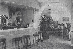 A grainy black-and-white newspaper photo of a restaurant interior.  To the left is a rounded bar with a barman behind and bar stools drawn up in front.  A large arch to the right leads through to a dining area with several seated parties.  The decor appears to be light and bright.