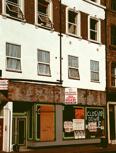 The ground, first, and second floors of a terraced double-width shop.  One of the ground-floor windows is boarded over, and the others have flyposters on.  “Closing down sale” has been spraypainted in one window, partially obscured by a flyposter. Windows on the upper floors are open, indicating those floors are still inhabited.