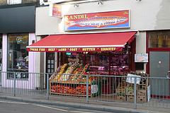 A terraced shopfront with vegetables including yams, peppers, chillies, and sacks of onions displayed at the front.  A sign above reads “Kandil / African Caribbean Food Land / Quality Meat & Fresh Fish”.  A red canopy stretches over the vegetables, with “Fresh Fish / Beef / Lamb / Goat / Mutten [sic] / Grocery” printed on it.