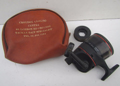 A brown leather bag with “Croydon Angling Centre / 65 London Rd Croydon / Tackle-Bait Specialist / Tel. 01-688 7564” printed on it in gold.  Next to it is a black fishing reel with red bands, around the same size as the bag.