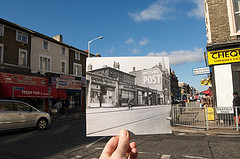 A vintage black-and-white photo held up in front of the same scene today.  The older shopfronts include Victor Value and Egleton Bros Ltd, while the modern ones include MBM Fresh Fish, Perfect Chicken, and Chat Patta.