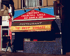 A terraced shopfront with a person walking past.  The front is covered with small, square, blue and blue-black tiles.  A blue and red sign above reads “The Croydon Charcoal Grill / Best Kebabs In Town”.