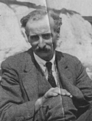 A black-and-white photo of a white man with receding wavy hair and a relatively substantial moustache.  He is wearing a suit with waistcoat and a tie, and appears to be sitting on the ground with a hand on one knee.  In the background is something that could be a rock or cliff.