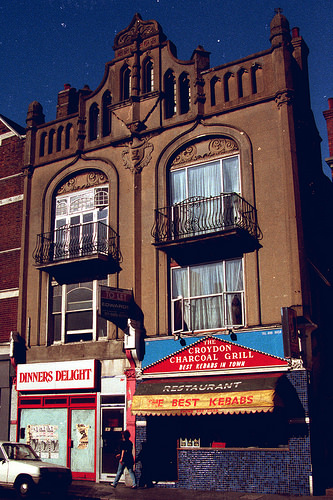 A view of all three storeys of two adjoining terraced addresses.  The shopfronts at ground level show “Dinners Delight” with whitewashed windows on the left and “The Croydon Charcoal Grill” on the right.  The second-floor windows have Juliet balconies and ornate arched tops.  A similarly ornate cornice is above.