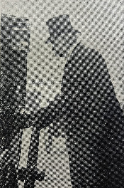 A black-and-white photo of a white man in a top hat and dark coat, seen sideways on, with his hand on the door of a carriage. He appears not to have noticed the photographer.