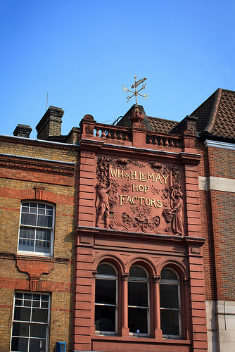 The upper floors of a narrow terraced building, seen in full sunlight.  The building is terracotta in colour, and has a sign on the top floor reading “WH & H LeMay Hop Factors” with an elaborately moulded surround.