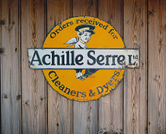A circular yellow/orange sign with a drawing of a person with pointed ears, wings, and a flat uniform cap.  A white bar runs across the centre of the sign with the words “Achille Serre Ltd.”, supplemented by curved wording on the circular part of the sign: “Orders received for [white bar here] Cleaners & Dyers”.