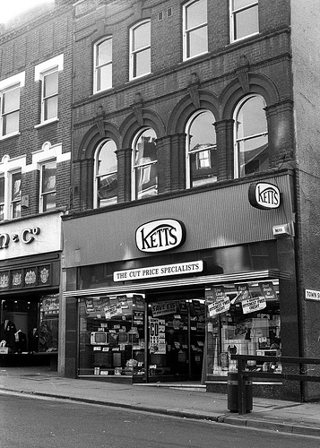 A black-and-white photo of an end-of-terrace shopfront. Signage above the ground-floor shop reads “Ketts / The Cut Price Specialists”.  Televisions, washing machines, and other electrical goods are visible in the window.