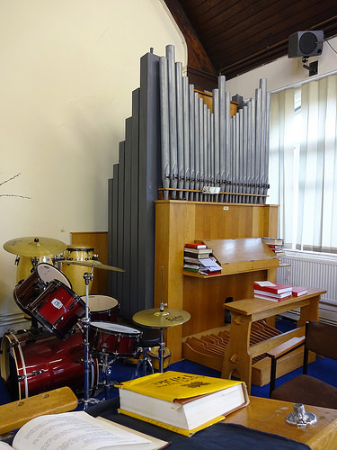 A colour photo of a small pipe organ in the corner of a room next to a drumkit.  A yellow bible is visible on a lectern in the foreground.