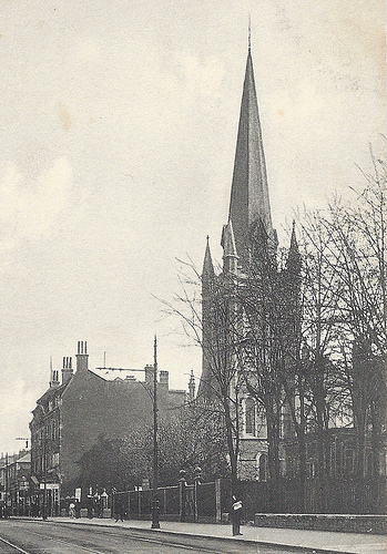 A black-and-white photo of a church with a spire towering over the other buildings around it.  Leafless trees stand in front, and tram tracks are just visible along the road.