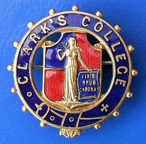 A round enamel badge in blue and red.  “Clark’s College” is embossed around the circumference, and a figure holding a sign reading “Finis Opus Coronat” is in the centre.