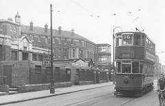 Black-and-white photo of an old-fashioned double-decker tram running along a road next to a tall, bulky building.  A sign on a post reads “Hospital / Please Drive Quietly”.  A cyclist is just visible in the foreground on the other side of the tram.